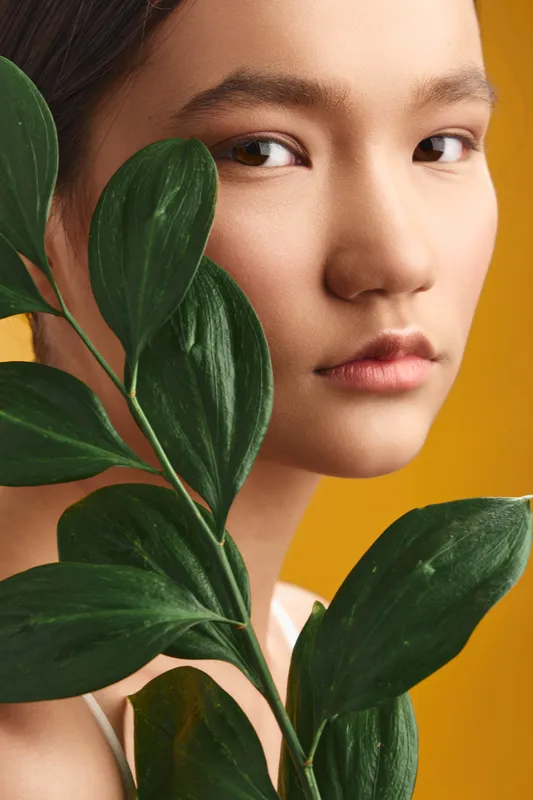 Asian woman with lush leaves held near her face stands in a vibrant studio setting, exuding calm confidence and contemporary style against a bright yellow backdrop.
