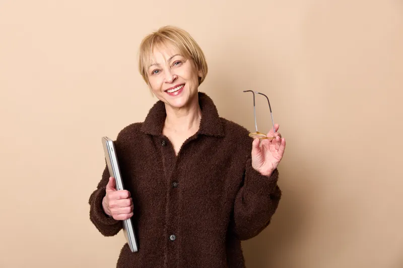 Elderly woman, smiling, portrait in studio, holding book and glasses, relaxed posture, warm beige background, soft light, casual learning mood, bright smile, looking at camera