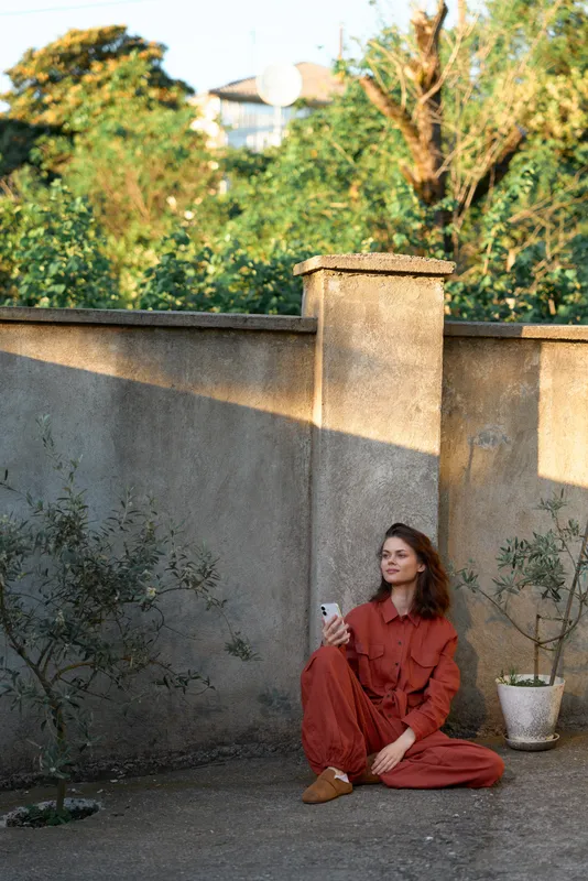 Young woman in rust red outfit sits on the ground beside a concrete wall gazing softly into the distance with a calm mood and relaxed pose