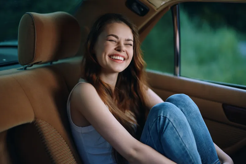 Happy young woman sitting in a car, enjoying a carefree moment, wearing a casual outfit, with a bright smile and a natural background highlighting positivity and freedom.