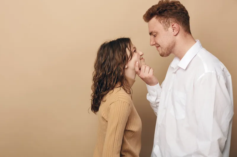 Young couple in casual clothes share a tender moment, smiling and looking into each other's eyes on beige background. Concept of love, affection, and relationship intimacy.