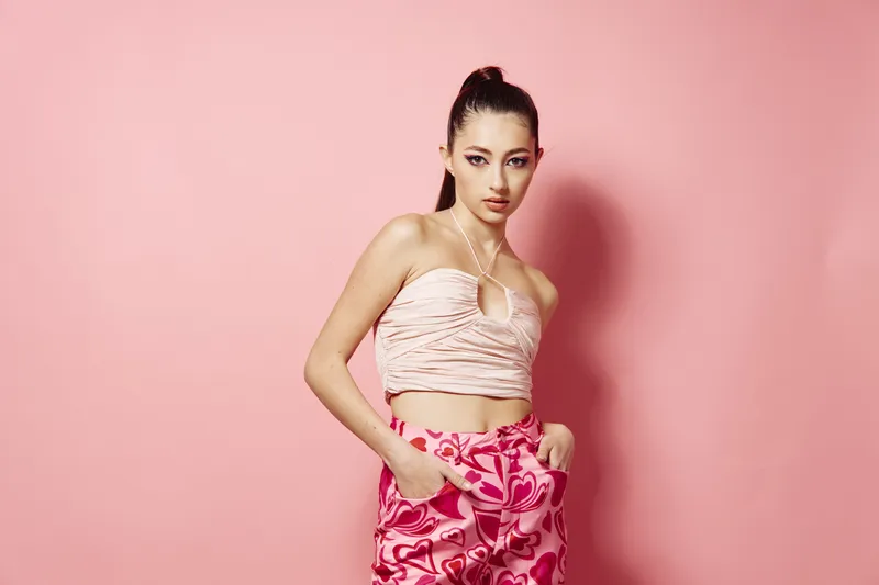 Young woman in a fashionable floral pants and a stylish ruched sleeveless top poses confidently against a pastel pink backdrop, embodying beauty, fashion, and creative style.