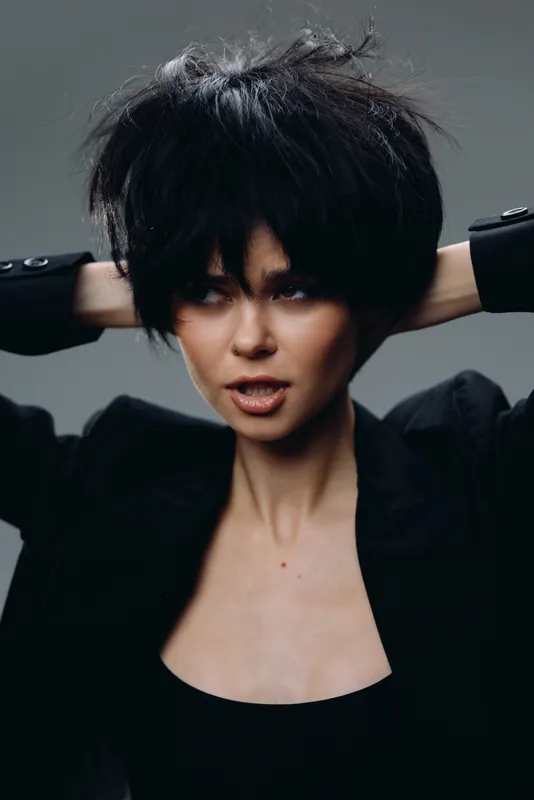 Young woman with short black hair wearing a black jacket posing confidently against a dark background