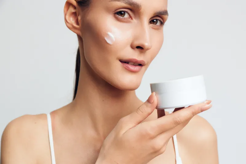 Young woman with long brunette hair applies white face cream on her cheek while holding a cosmetic jar against a plain light background, showing skincare routine and natural beauty.