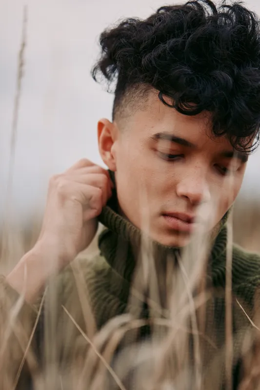 Close portrait of a young man in tall grass wearing a turtleneck sweater, capturing natural facial detail and quiet emotion that reflects authenticity and veracity in a candid moment.