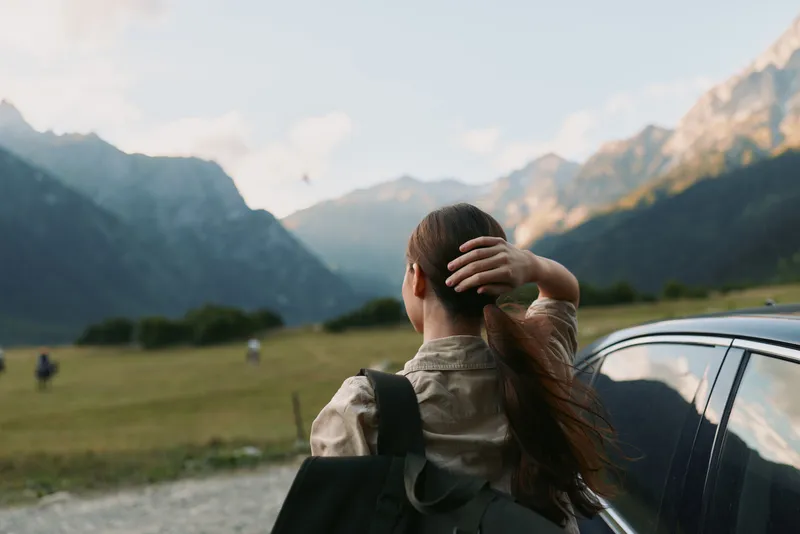 Woman lifestyle traveler standing near car looking at mountain landscape with hand in hair, casual outfit, peaceful moment, outdoor adventure, natural scenery