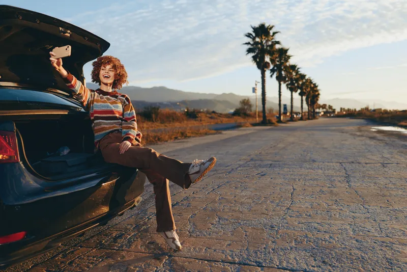 Woman lifestyle outdoors by car trunk on a quiet road, warm sunlight desert air, relaxed pose with legs out, casual striped shirt and jeans, palm trees and distant mountains scenic background, travel