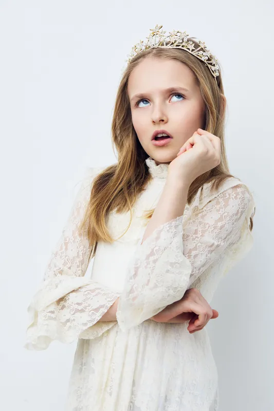 Elegant young girl in a lace dress with a tiara, expressing curiosity against a minimalist white background, perfect for children s fashion and portrait photography