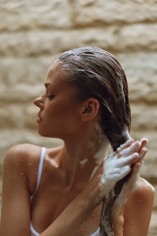 Woman washing hair with foam shampoo, wet skin, natural light, spa relaxation, personal care, calm mood indoors.