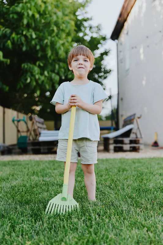 Happy boy holding a rake in a sunny garden, wearing casual clothes, enjoying outdoor activities and helping in the yard, showcasing a joyful summer vibe.