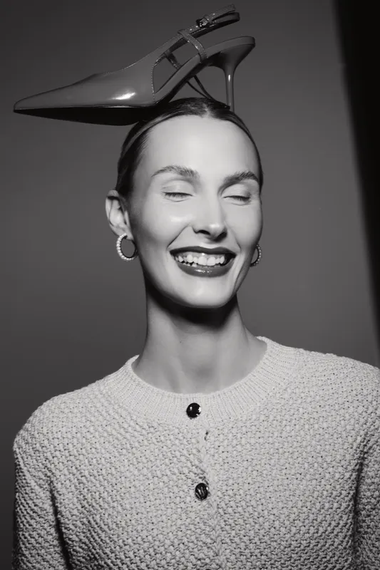 Smiling white woman with pale skin and slicked back hair balances high heel shoe on head in black and white studio portrait. Concept of fashion and playful elegance.