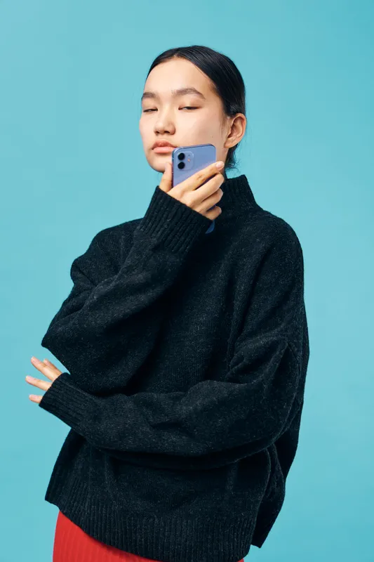 Young woman in black sweater holding smartphone near face with closed eyes against blue background for technology, connection, communication and modern lifestyle concept.
