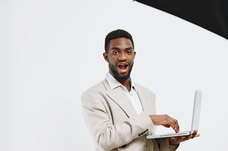 Smiling man with dark skin and short black hair holding a silver laptop in hand, wearing a beige sweater and white shirt, look camera, studio portrait white background positive emotion. People lifesty