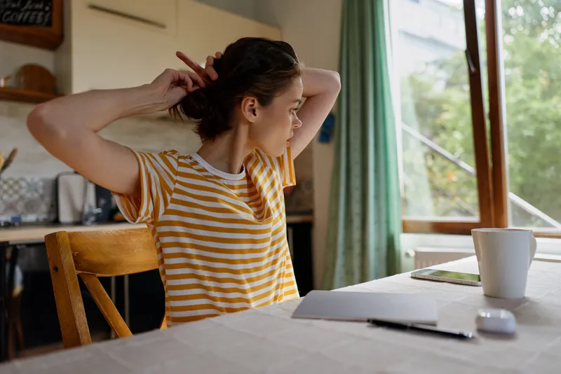 A woman in a striped shirt sits at a bright kitchen table, tying her hair as sunlight spills through a window, capturing a simple morning routine and home selfcare portrait.