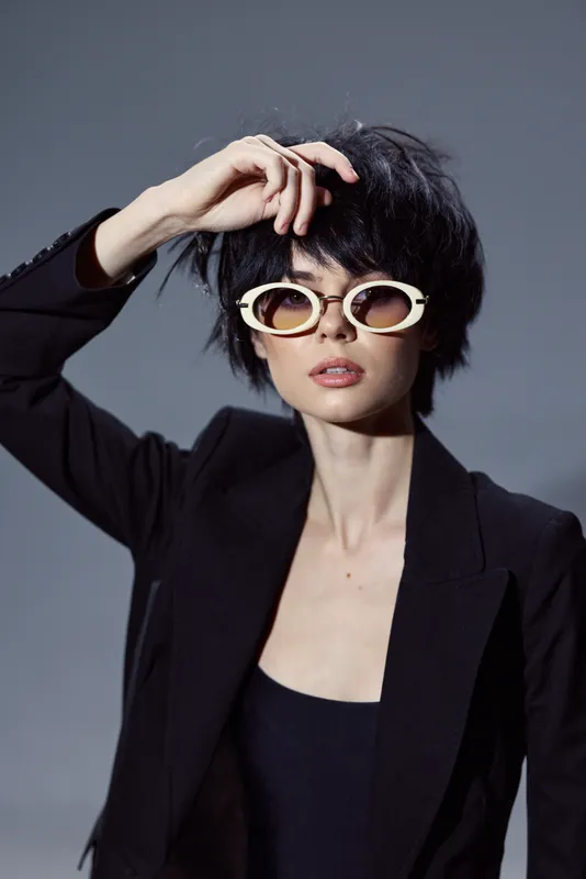 Stylish woman wearing black blazer and white sunglasses posing confidently with hand on head
