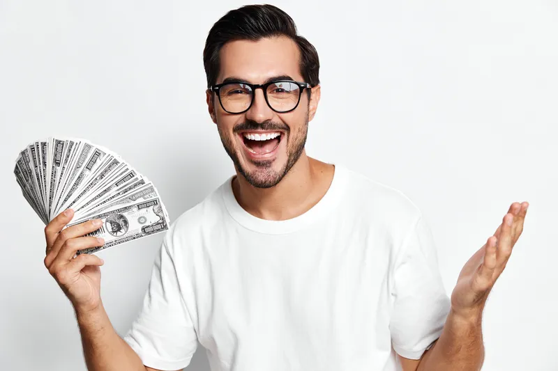 Happy smiling man holds fan of dollar bills, wearing glasses and white t-shirt, looks at camera with excitement against a light gray background. People finance concept.