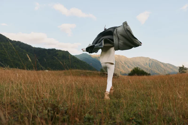 Young woman with long hair walks through a grassy field surrounded by mountains while holding a coat above her head in a carefree moment.