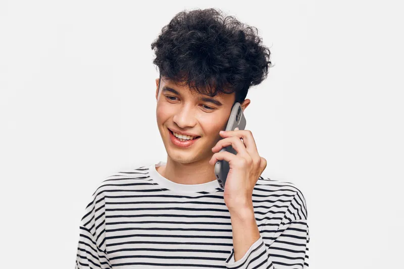 Young man talking on smartphone with joyful smile, wearing striped shirt against white background, casual style and positive mood in indoor studio setting.