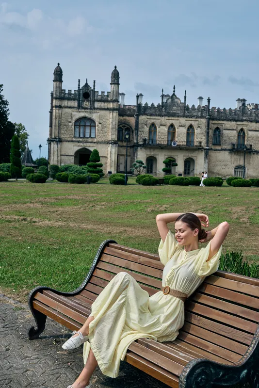A woman in a light dress lounges on a wooden bench in a manicured garden, with a grand historic castle in the background, creating a serene, stylish outdoor scene.