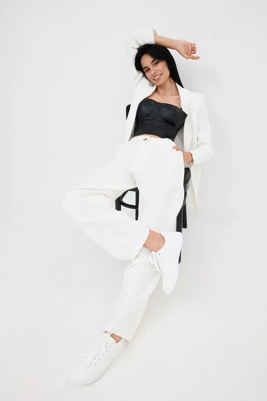 relaxed woman sitting on chair wearing white suit and black top with casual white sneakers against plain white background, modern fashion style, confident pose