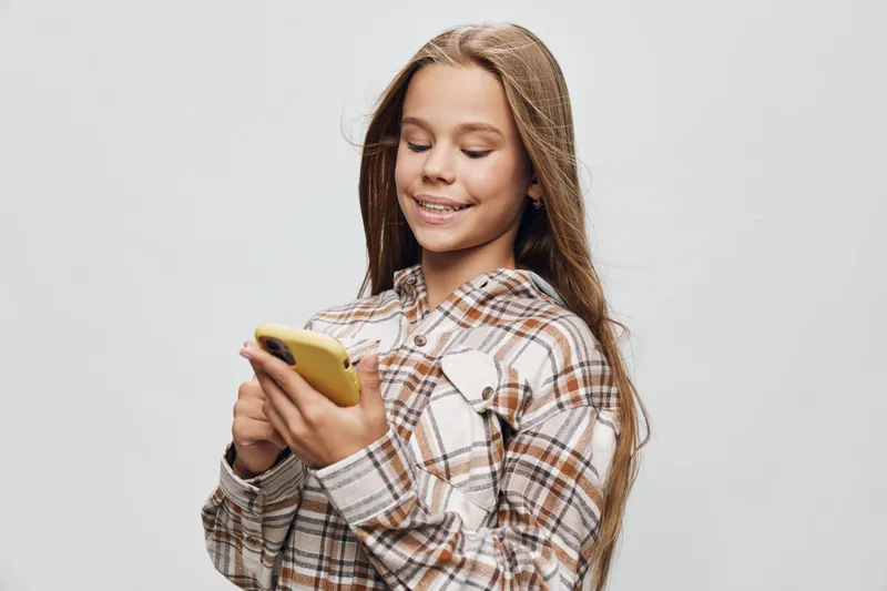 Girl smiling and texting on smartphone with braces wearing plaid shirt standing against white background. Happy teenager using mobile phone for communication and social media online.