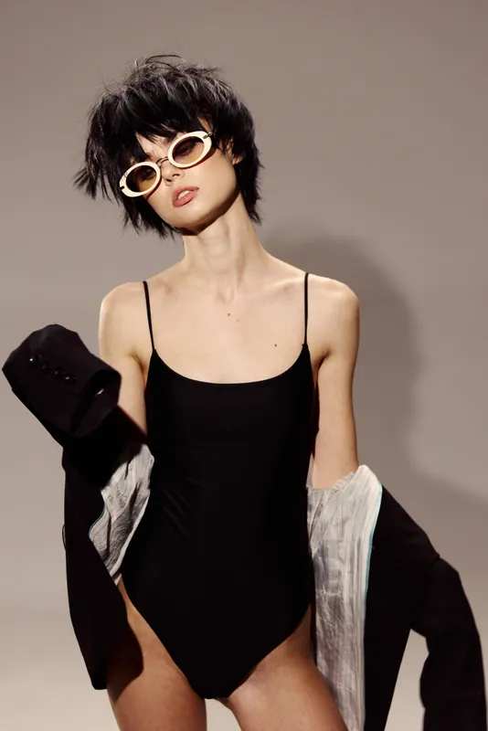 Stylish woman wearing black swimsuit and sunglasses posing with jacket in minimalist studio setting.