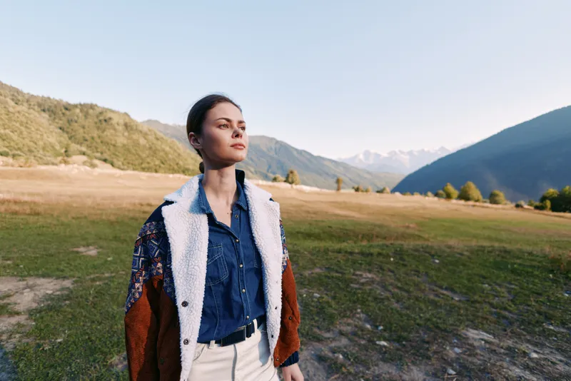 Woman portrait in mountains and nature on a valley field, wearing a jacket and denim shirt for travel and outdoors lifestyle, thoughtful pose with scenic horizon and sunlight.