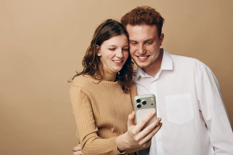 Young couple smiling and taking a selfie together with a smartphone. They stand close, showing affection and happiness in a casual indoor setting with neutral background.