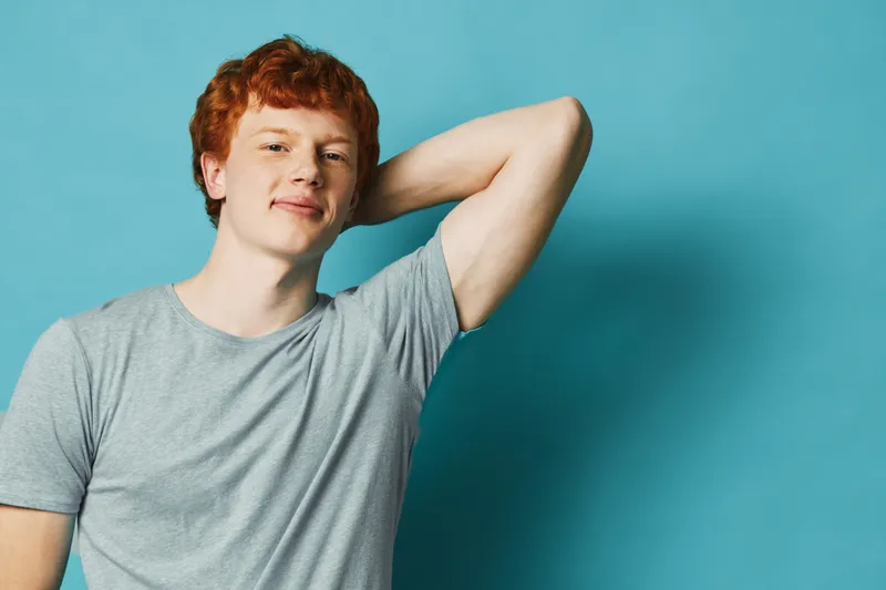 Young man with red hair in gray t shirt posing confidently with hand behind head against blue background studio portrait casual style and smile