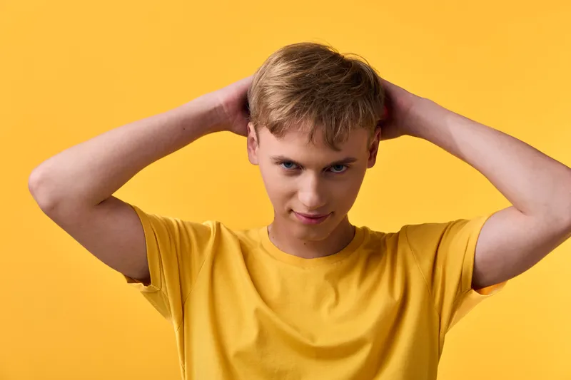 Boy with hands on head in a yellow shirt against a bright background, energetic and curious mood, casual and vibrant color in a simple setting