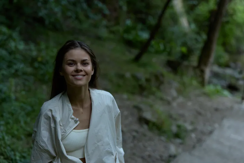 smiling woman outdoors wearing white jacket and top, surrounded by green forest with soft natural light and peaceful atmosphere during daytime