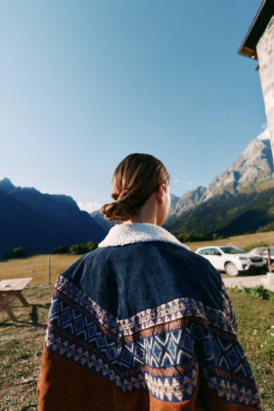 Woman back looking at mountains in the countryside, wearing a patterned jacket. Outdoors travel scene with parked car, alpine valley, rural house and clear summer sky.