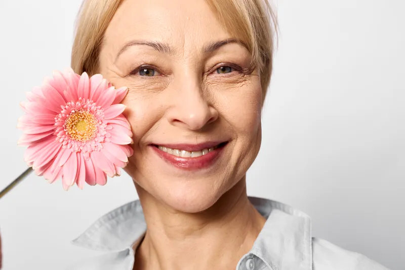 Smiling senior woman in healthcare scrubs nurse holding a pink gerbera flower to her cheek in a clean studio portrait, conveying calm warmth and kindness