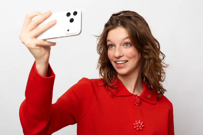 Young woman in red sweater takes a selfie with a smartphone, smiling and looking excited. White background highlights casual lifestyle and modern technology use in everyday moments.