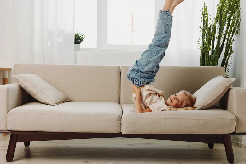 Happy child playing on beige sofa in bright living room, lifting legs in air and smiling. Cozy home interior with cushions and plants in natural light. Concept for childhood joy and playtime.