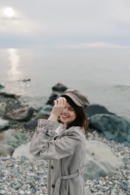 Smiling woman in checkered coat and hat stands on rocky beach near sea during cloudy day. Concept for fashion, autumn style, outdoor lifestyle, and natural beauty with serene ocean background.