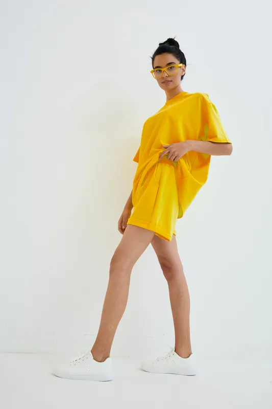 Young woman wearing bright yellow casual outfit and white sneakers posing confidently against white background, showcasing modern street style fashion and cool attitude.