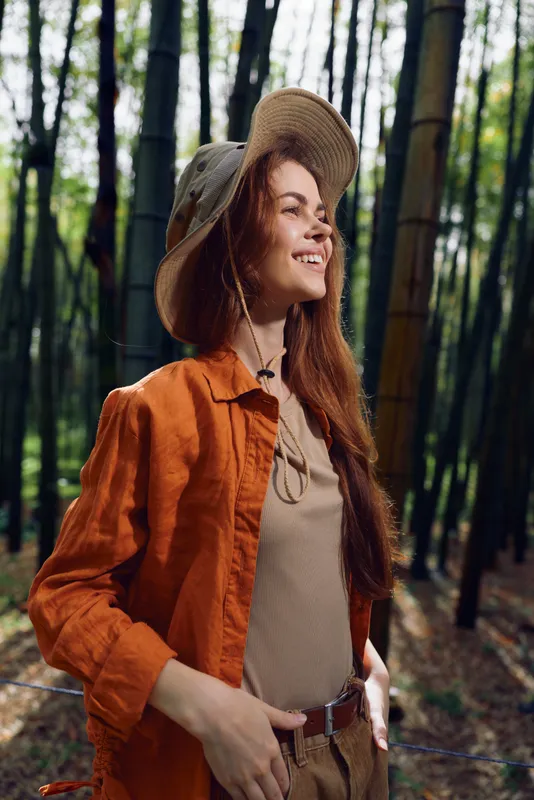 Woman smiling in a bamboo forest wearing a straw hat and orange jacket, long hair flowing, hands on hips, enjoying nature light and a peaceful outdoor portrait in green woodland.