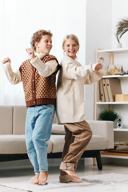 Two cheerful children dance indoors wearing cozy sweaters, barefoot on a soft rug, exuding playful energy and warm holiday vibe in a bright living room.