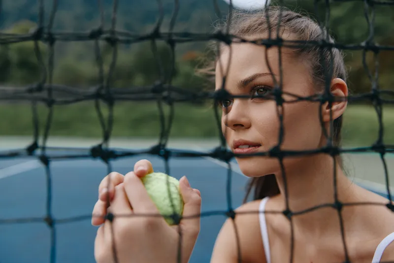 Female tennis player holding a ball with a focused expression, surrounded by a tennis net in a vibrant outdoor setting, showcasing athleticism and concentration