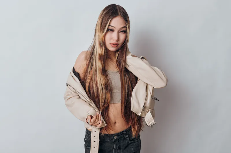 Young woman with long hair wearing cropped top and jacket posing in studio. Casual fashion model, standing against white background with confident expression and stylish look.