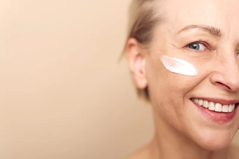 Close-up of smiling mature woman applying white face cream on cheek, natural skincare concept with soft light on beige background showing healthy skin and beauty care routine.