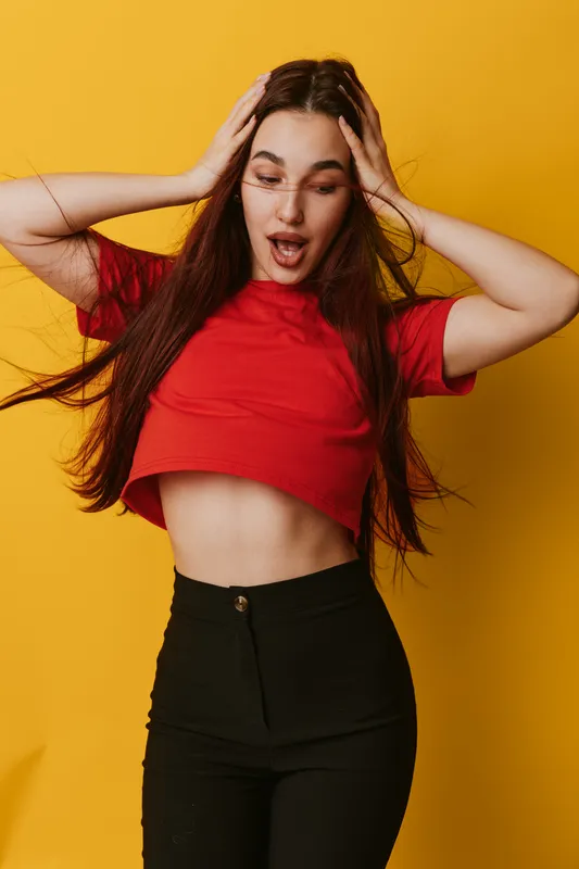 Young woman with long hair in red cropped shirt and black pants posing surprised with hands on head on yellow background. Female model expressing shock and excitement indoors.