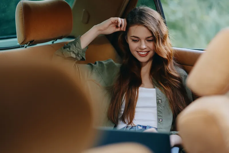Young woman with long brown hair smiling while using a laptop in a car, casual outfit features a green jacket on a sunny day, conveying a sense of relaxation and happiness.