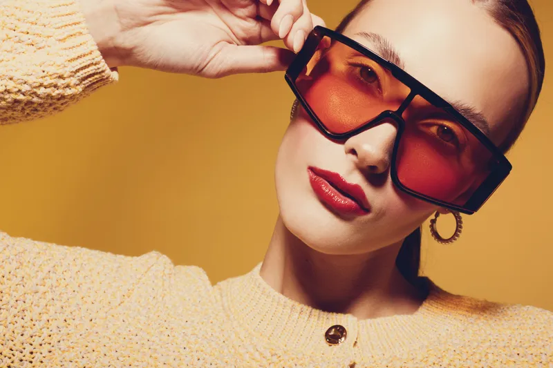 Stylish woman in oversized orange sunglasses and yellow sweater poses against orange background. Fashion portrait with bold makeup and confident expression. Concept of modern style and accessories.