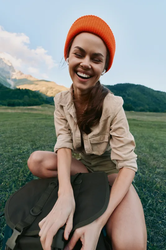Young woman with long brown hair wearing an orange beanie smiles joyfully while sitting outdoors on a backpack in a green mountainous landscape under blue sky.