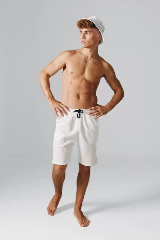 Young man standing barefoot in white shorts and bucket hat looking sideways with hands on hips against plain background in studio.