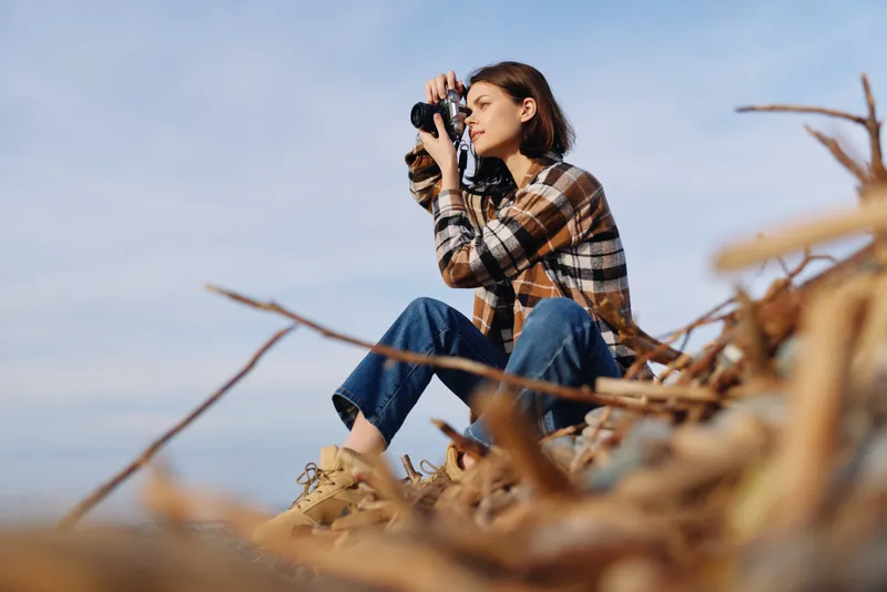 Female photographer perched among dry branches, kneeling in a casual plaid shirt and jeans, capturing coastal scenery with a compact camera under a clear blue sky, and warm light.