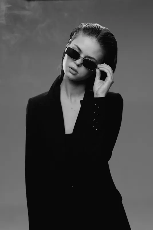 Black and white portrait of a stylish young woman wearing dark sunglasses and a black blazer with a confident expression.