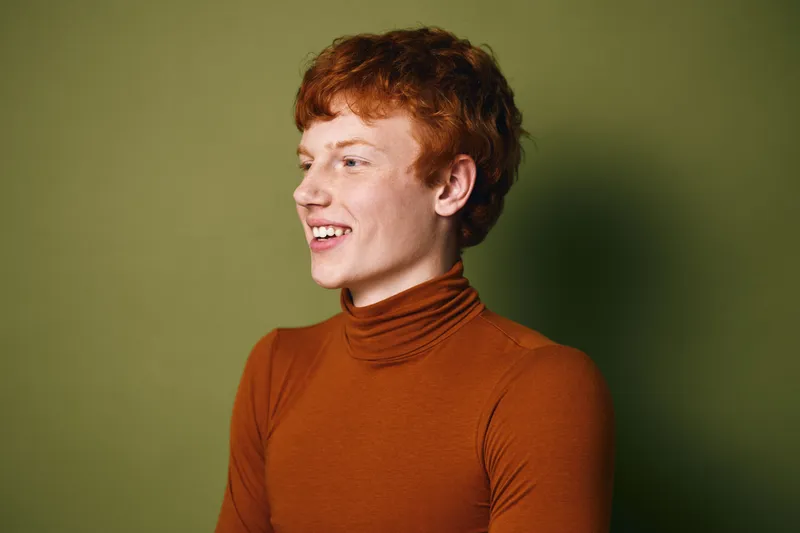 Young redhead person in orange turtleneck smiling looking to side against green background, natural light portrait with casual style and happy expression.